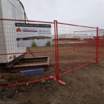 Red temporary welded wire mesh fence with flat base feet, securing a dirt construction site. A CANSUPPLY wholesale sign is attached to the fence.