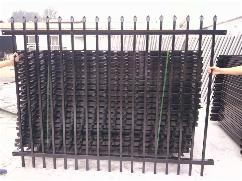 Two people holding up a black steel spear-top fence panel in front of a large, strapped stack of identical panels.