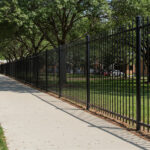 Long black metal picket fence running alongside a sidewalk, providing perimeter security for public grounds or a school.