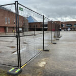 Black temporary mesh fencing with green and yellow bases enclosing an area on a concrete tarmac next to a brick building, possibly at an airport, under an overcast sky.