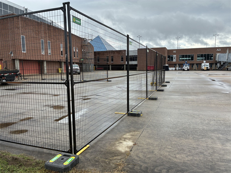 Black temporary mesh fencing with green and yellow bases enclosing an area on a concrete tarmac next to a brick building, possibly at an airport, under an overcast sky.