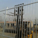 A close-up of a galvanized steel picket fence with a forklift in the industrial background.