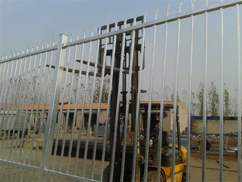 A close-up of a galvanized steel picket fence with a forklift in the industrial background.