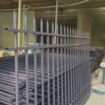 Unpainted steel picket fence panels with pressed spear tops are stacked inside a manufacturing facility before the final coating process.