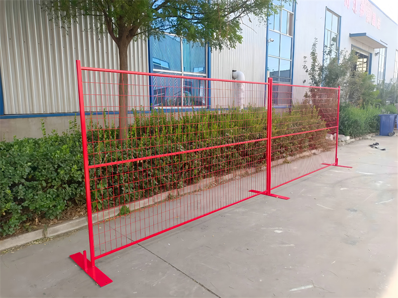 Two connected red temporary mesh fence panels with red T-shaped bases, displayed on a concrete pad with green foliage and a building in the background.