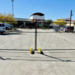 Black temporary mesh fencing with yellow bases enclosing an area in a paved parking lot, with vehicles and a building in the background.