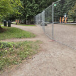 A long line of galvanized temporary mesh fencing with red bases, curving along a dirt path in a park-like setting with green trees and grass.