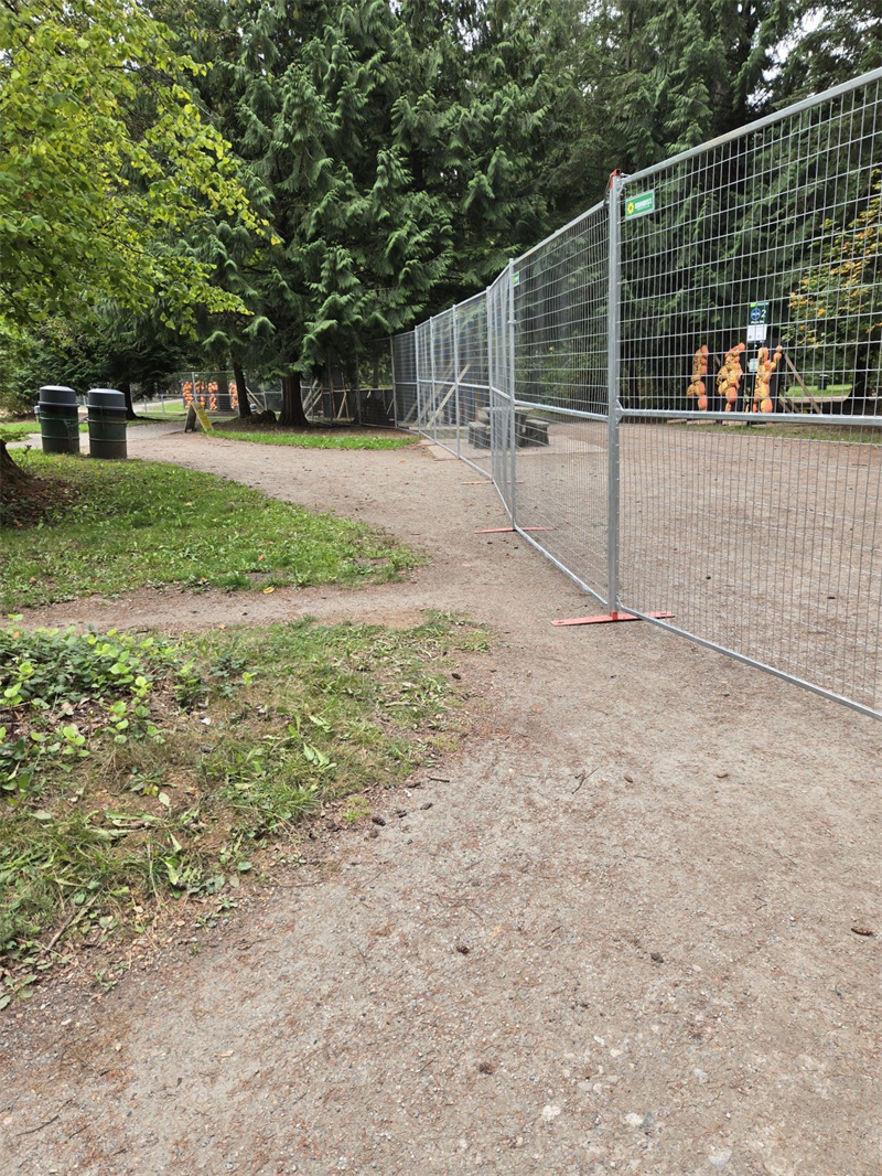 A long line of galvanized temporary mesh fencing with red bases, curving along a dirt path in a park-like setting with green trees and grass.