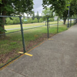 Dark green temporary fencing with yellow plastic bases lines a paved sidewalk, separating it from a grassy park area with trees.