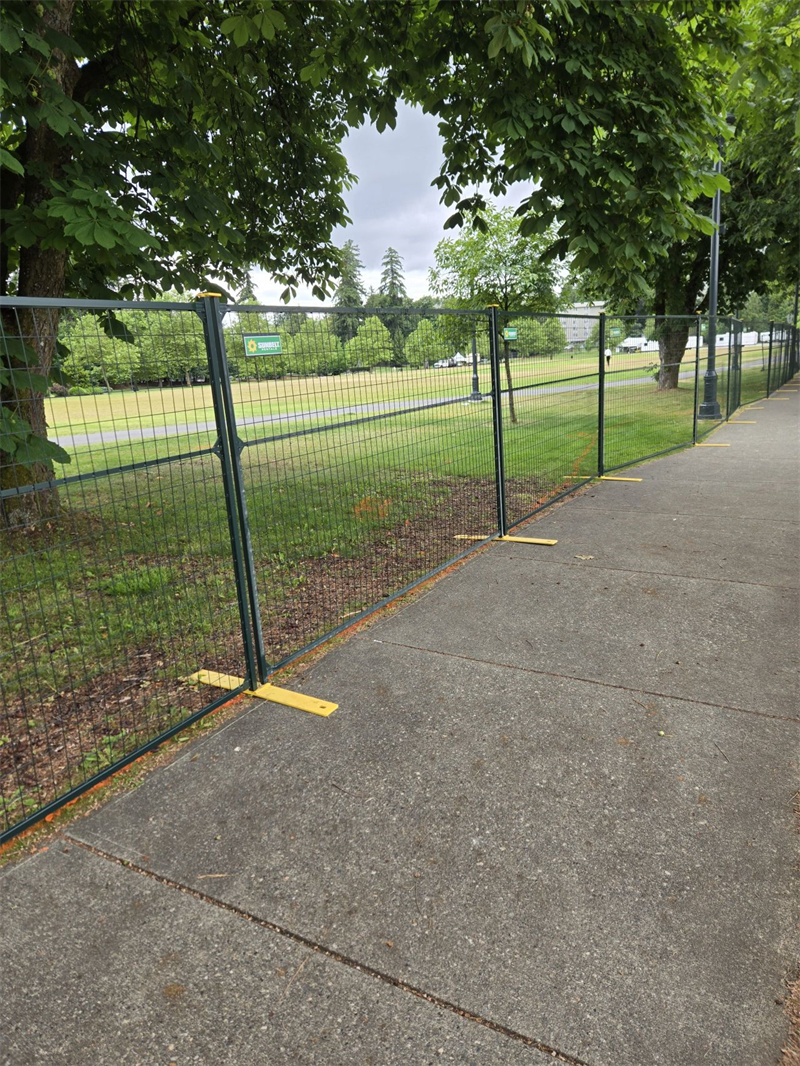 Dark green temporary fencing with yellow plastic bases lines a paved sidewalk, separating it from a grassy park area with trees.