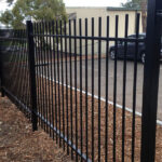 A tall, black flat-top steel security fence installed along the perimeter of a commercial property parking lot.