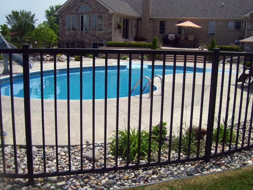 A black vertical picket fence surrounding an in-ground swimming pool in a landscaped residential backyard, with a large house visible in the background.
