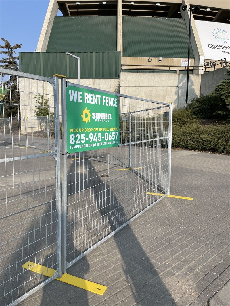 Galvanized temporary fencing with yellow bases prominently displays a green "WE RENT FENCE" sign from Sunbelt Rentals, set against a stadium structure.