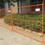 Orange temporary fence panels with bases are set up on a paved area next to green bushes and a building, providing a temporary barrier.
