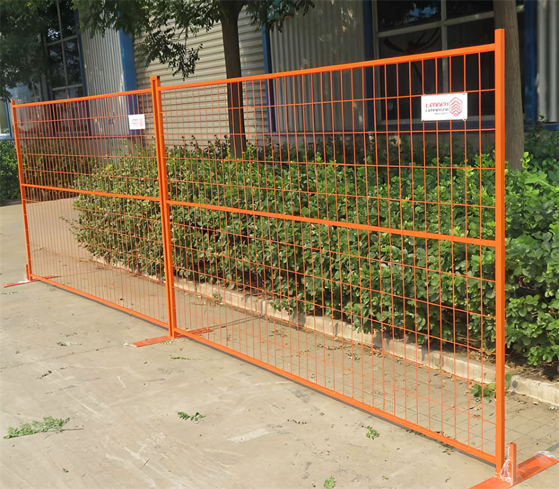 Orange temporary fence panels with bases are set up on a paved area next to green bushes and a building, providing a temporary barrier.