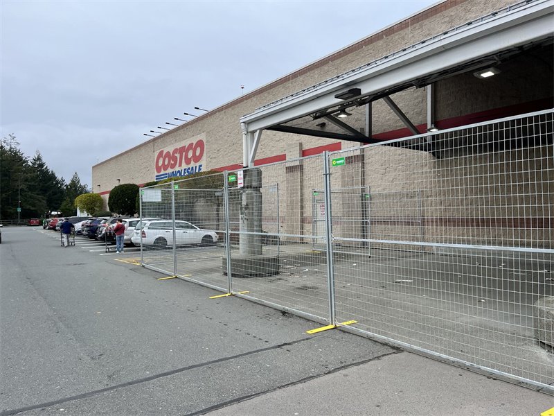 Galvanized temporary fencing with yellow plastic bases in front of a Costco Wholesale store, separating the parking lot from the building entrance.