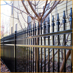 A black ornamental steel fence with decorative finials installed in front of a house with beige siding and a bare tree.