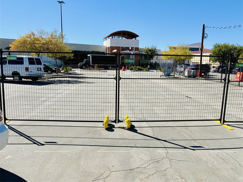 Black temporary fence panels with yellow plastic bases securing an outdoor area, possibly a construction site or event space. A white van and other vehicles are visible in the background, along with a building and trees.