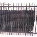 Two people holding up a black steel spear-top fence panel in front of a large, strapped stack of identical panels.