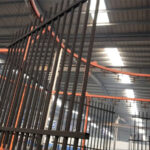 A steel fence panel hangs from an overhead conveyor system as it moves through a factory's automated finishing line, highlighting a key stage in the quality coating process.