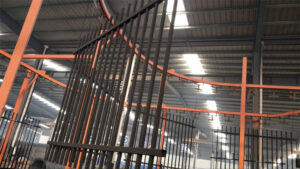 A steel fence panel hangs from an overhead conveyor system as it moves through a factory's automated finishing line, highlighting a key stage in the quality coating process.