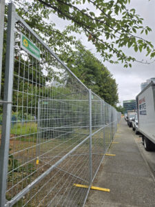 A long line of galvanized temporary fencing with yellow plastic bases secures the perimeter of a construction site, backed by lush green trees and foliage.