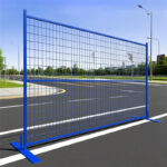 A single blue temporary welded wire mesh fence panel with flat bases, standing on a paved road with a curb and green space in the background.