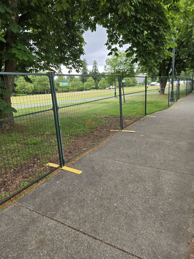 Dark green temporary fencing with yellow plastic bases lines a paved sidewalk, separating it from a grassy park area with trees.