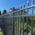 A close-up, low-angle shot of a black aluminum or steel picket fence with decorative, pointed finials. The fence is set against a bright blue sky, with lush green bushes and the roof of a house visible in the background.