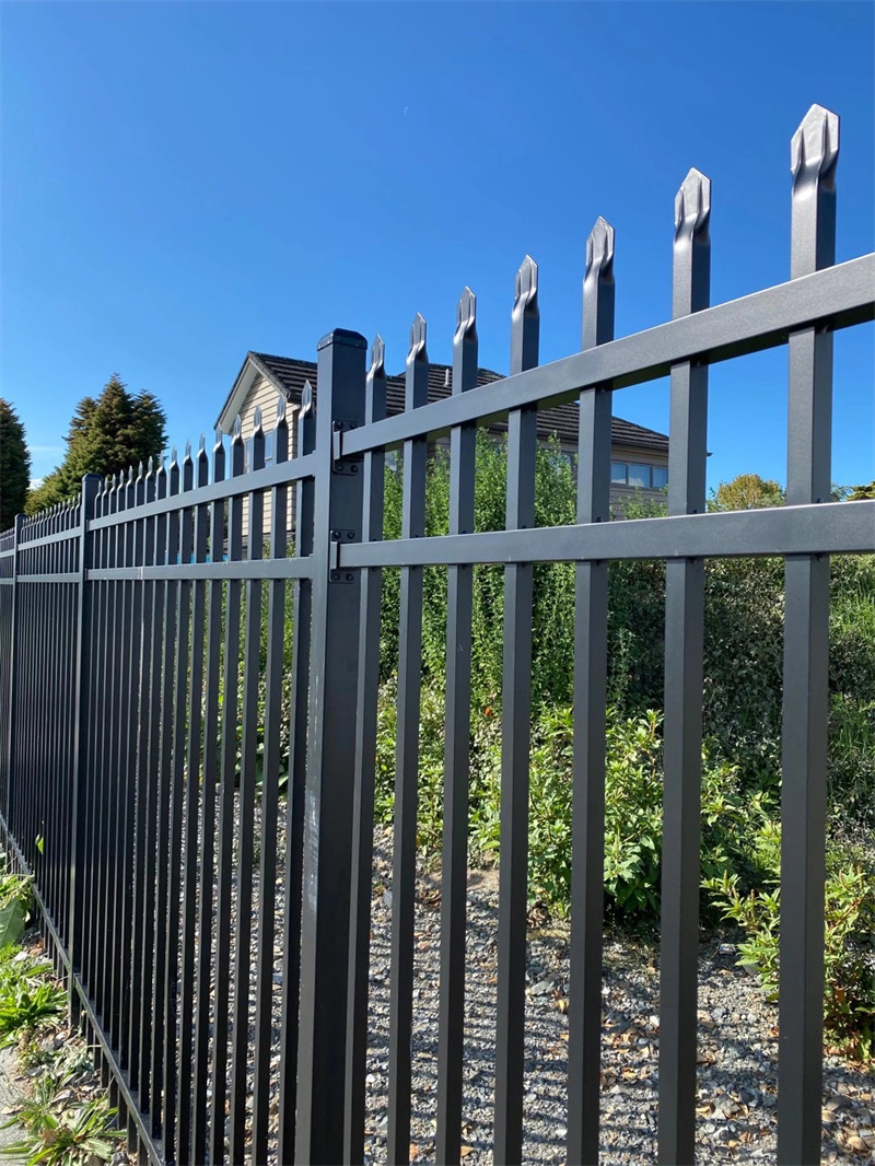A close-up, low-angle shot of a black aluminum or steel picket fence with decorative, pointed finials. The fence is set against a bright blue sky, with lush green bushes and the roof of a house visible in the background.
