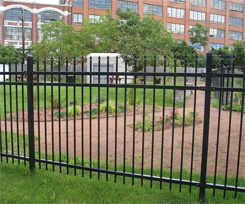 A black steel picket fence enclosing a public park with a grassy area and a gravel path. In the background, there are trees and a large, multi-story brick industrial building.