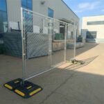 Close-up of BRAVO ballast installed on chain link fence post - detailed view of fence weight attachment with branded black base and yellow safety strips