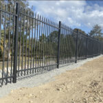 A long black steel security fence with spear tops installed on a gravel and dirt perimeter, with trees and a blue sky with clouds in the background.