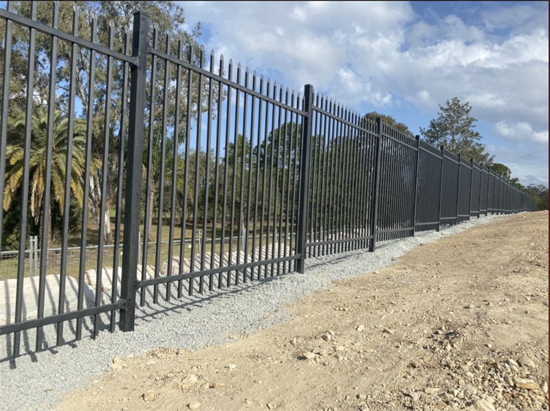 A long black steel security fence with spear tops installed on a gravel and dirt perimeter, with trees and a blue sky with clouds in the background.