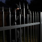 A person dressed in black clothing, including a beanie and gloves, attempting to climb a tall, black steel picket fence with pointed tops during the night. The person's face is partially visible as they grip the pickets.