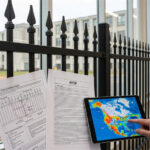 An image showing fence standards being reviewed. In the foreground, a person holds a tablet displaying a weather map of North America, with papers showing ASTM fence specifications resting in front of it. A black steel picket fence is visible in the background.