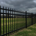 A long black steel picket fence with pointed tops stretching across a vast green field under a dark, cloudy sky. In the far background, houses and a line of trees can be seen.