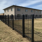 A photo of a black aluminum or steel fence surrounding the property of a large, two-story light-colored brick building. The fence is installed on a dormant, grassy lawn under a clear blue sky. A concrete sidewalk runs alongside part of the fence line.