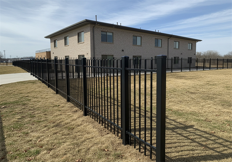 A photo of a black aluminum or steel fence surrounding the property of a large, two-story light-colored brick building. The fence is installed on a dormant, grassy lawn under a clear blue sky. A concrete sidewalk runs alongside part of the fence line.