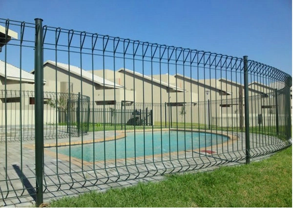Dark security fencing with a curved rolled top enclosing a kidney-shaped swimming pool, with single-storey houses and a lawn in front under a clear sky