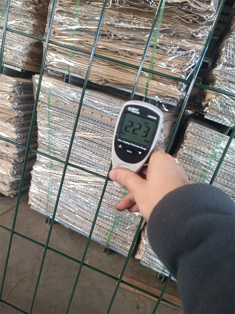 A hand holds a digital moisture meter in front of DB Fencing's green temporary fence, with stacked wire mesh panels wrapped for export in the background, highlighting the company's rigorous quality assurance for durable, galvanized fencing products.