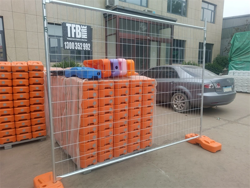 Temporary wire mesh fence with orange plastic feet, manufactured by DB Fencing (Anping Deban Metal Wire Mesh Products Co., Ltd), a leading one-stop fence supplier with in-house plastic feet production, displayed in a warehouse setting with stacked panels and rental signage.