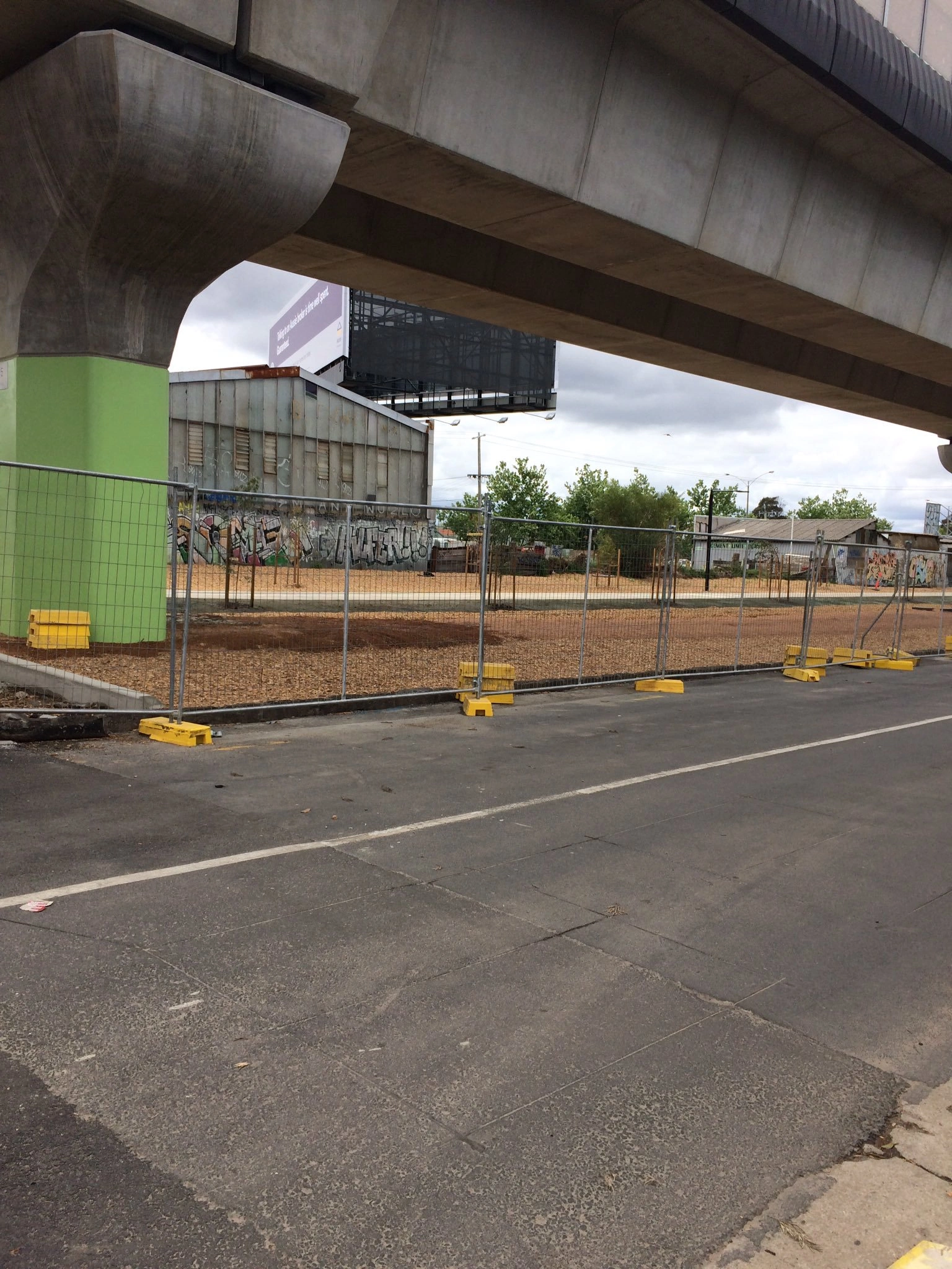 A temporary fencing system, featuring fence panels anchored by distinctive yellow plastic feet, secures a construction or development site under a concrete overpass in an urban Australian environment. Anping Deban Metal Wire Mesh Products Co., Ltd (DB Fencing) provides high-quality, AS 4687-2022 compliant Temporary Fencing Systems for Construction Sites, including these durable, in-house manufactured plastic feet, vital for Australian construction and civil engineering firms.