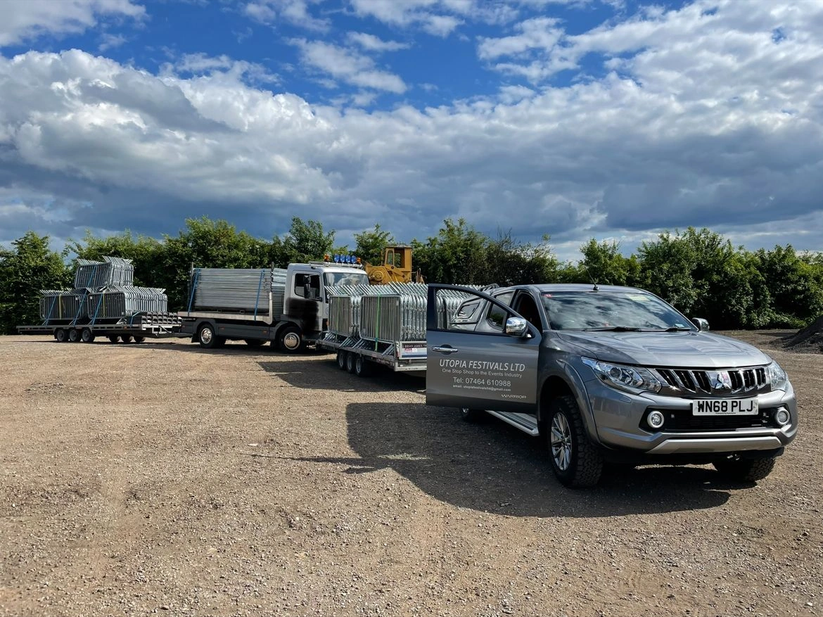 A fleet of vehicles, including a pickup truck branded for Utopia Festivals LTD and a lorry, efficiently transporting multiple stacks of durable crowd control barriers. This image showcases DB Fencing's high-quality, hot-dipped galvanized crowd control barriers being prepared for large events and public gatherings, emphasizing our robust logistics for event management companies.