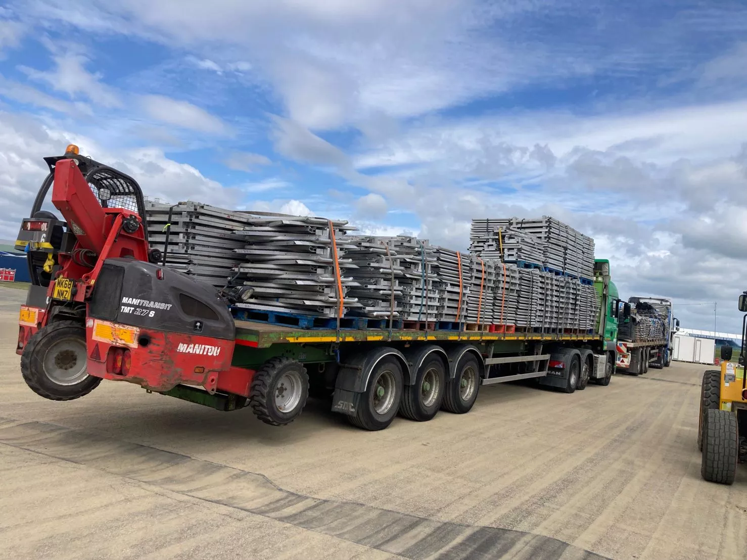A red Manitou telehandler loads stacks of galvanized Crowd Control Barriers & Event Perimeter Management components onto a flatbed truck, illustrating DB Fencing's large-scale manufacturing and global export logistics for B2B clients worldwide.