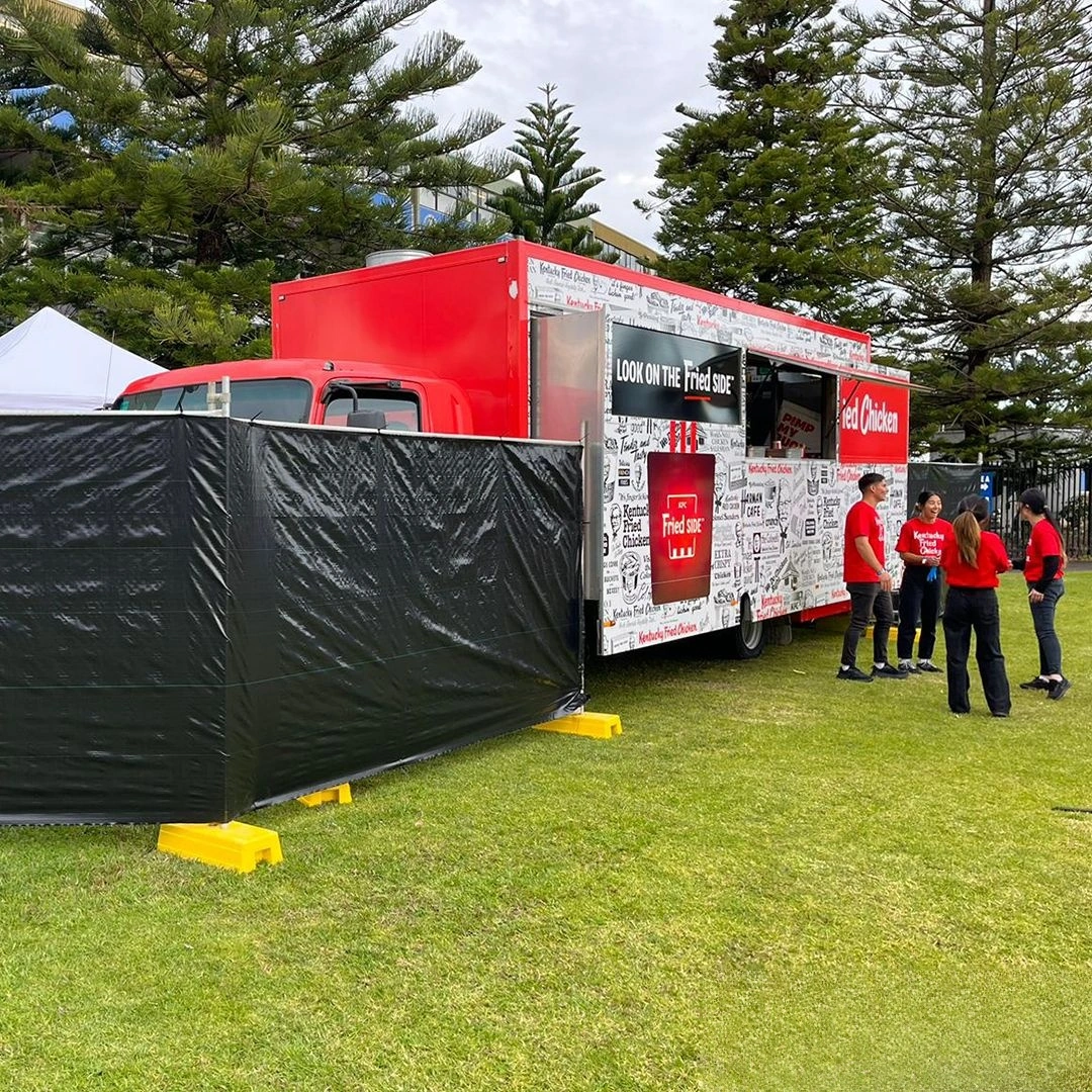 Robust DB Fencing crowd control barriers are deployed with black temporary fence panels and bright yellow plastic feet, ensuring public safety and efficient crowd management around a bustling food truck at an outdoor event. As a leading manufacturer, DB Fencing supplies event management companies with durable and compliant temporary fencing systems, including essential fence feet and base systems, designed for quick setup and stability in various environments.