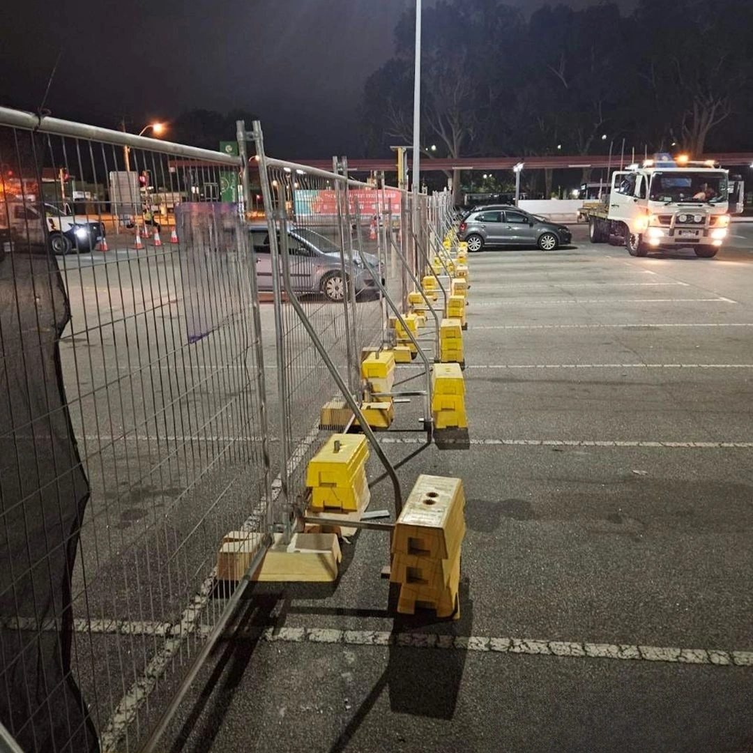 A long line of DB Fencing's signature yellow plastic temporary fence feet supporting galvanized temporary fencing panels, illuminated at night in a large parking area. This visual emphasizes our robust Temporary Fence Feet & Base Systems, manufactured in Anping, China, and designed to provide stable, compliant perimeters for construction sites or events, meeting Australian standards for global export.
