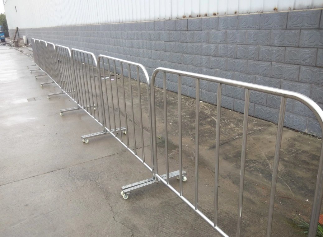 A series of shiny, portable steel crowd control barriers, manufactured by DB Fencing, are lined up on a wet concrete surface next to an industrial building. These durable crowd control barriers feature wheeled bases for easy deployment, showcasing DB Fencing's high-quality solutions for event management companies and distributors requiring reliable public safety and perimeter control.