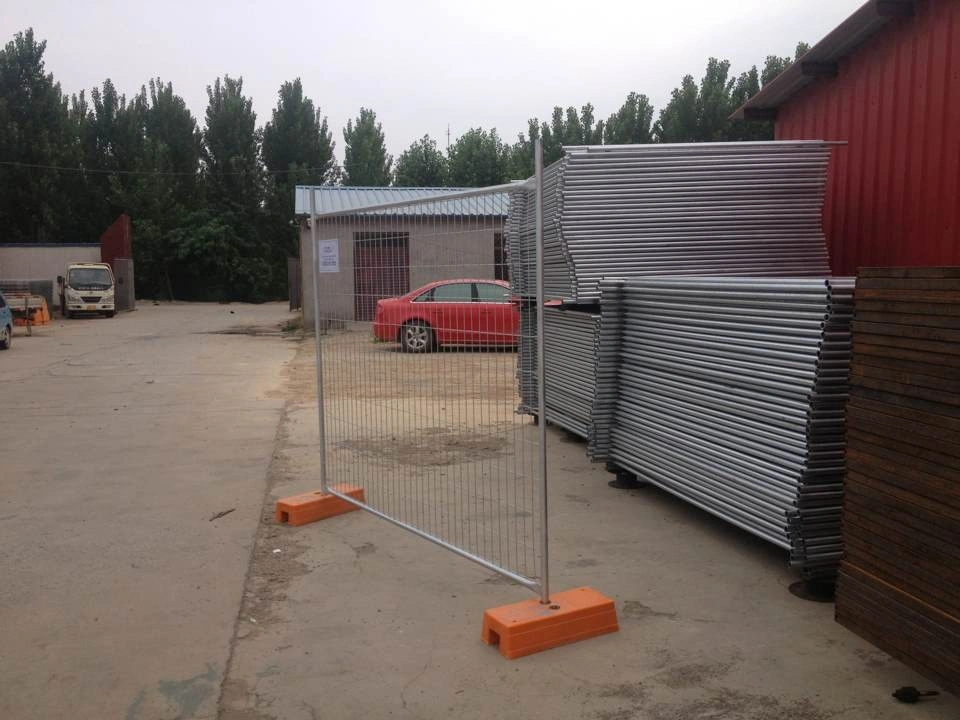 A galvanized temporary fencing system, featuring DB Fencing's proprietary orange plastic feet, is displayed in an industrial yard alongside stacks of fence tubes. This image represents Anping Deban Metal Wire Mesh Products Co., Ltd.'s high-quality Temporary Fencing Systems for Construction Sites, manufactured for durability and compliance with export standards, especially for markets like Australia.