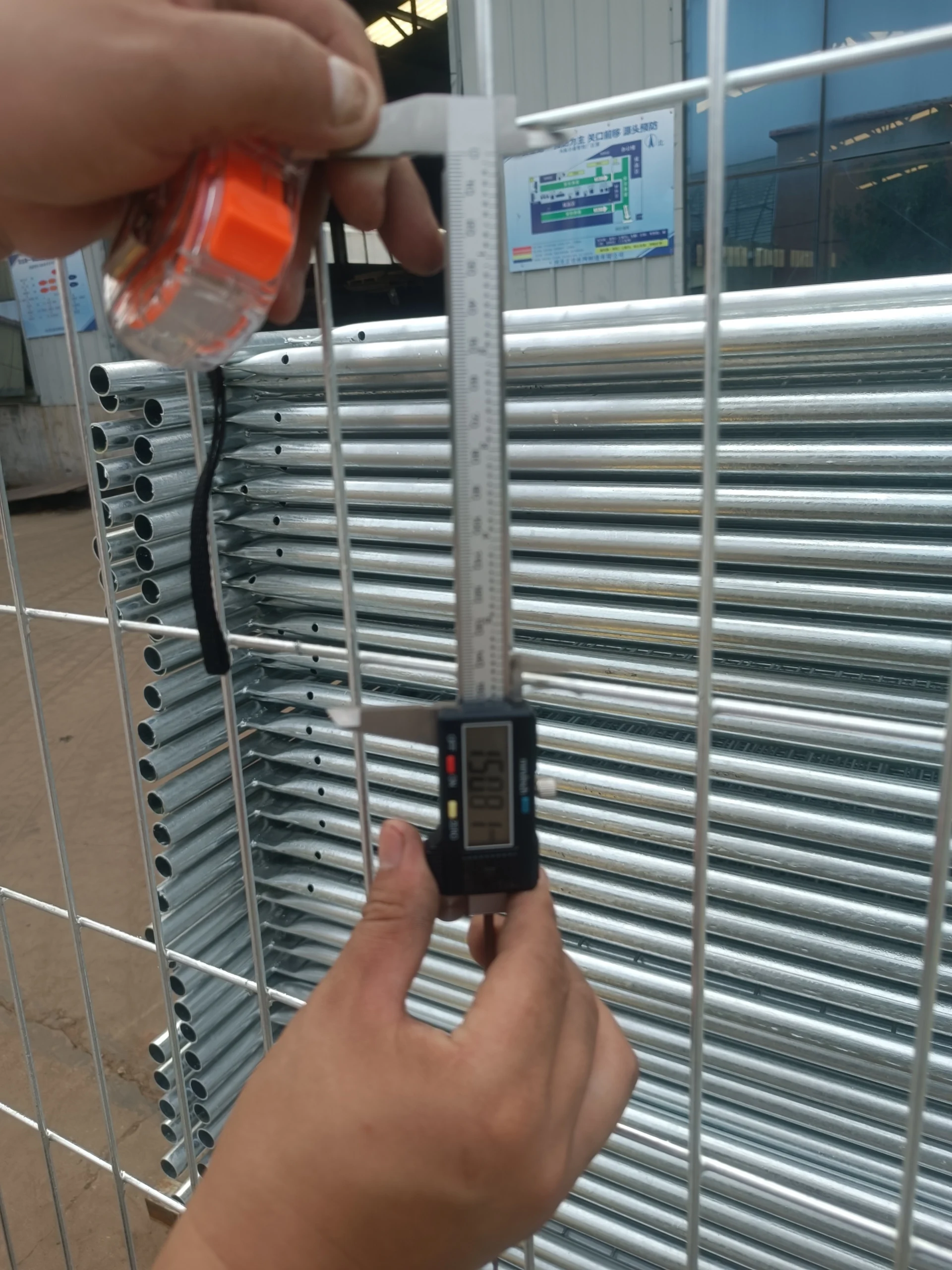 Quality control technician measuring a galvanised steel tube with a digital caliper against a stack of tubing, with a tape measure and factory wall signage behind